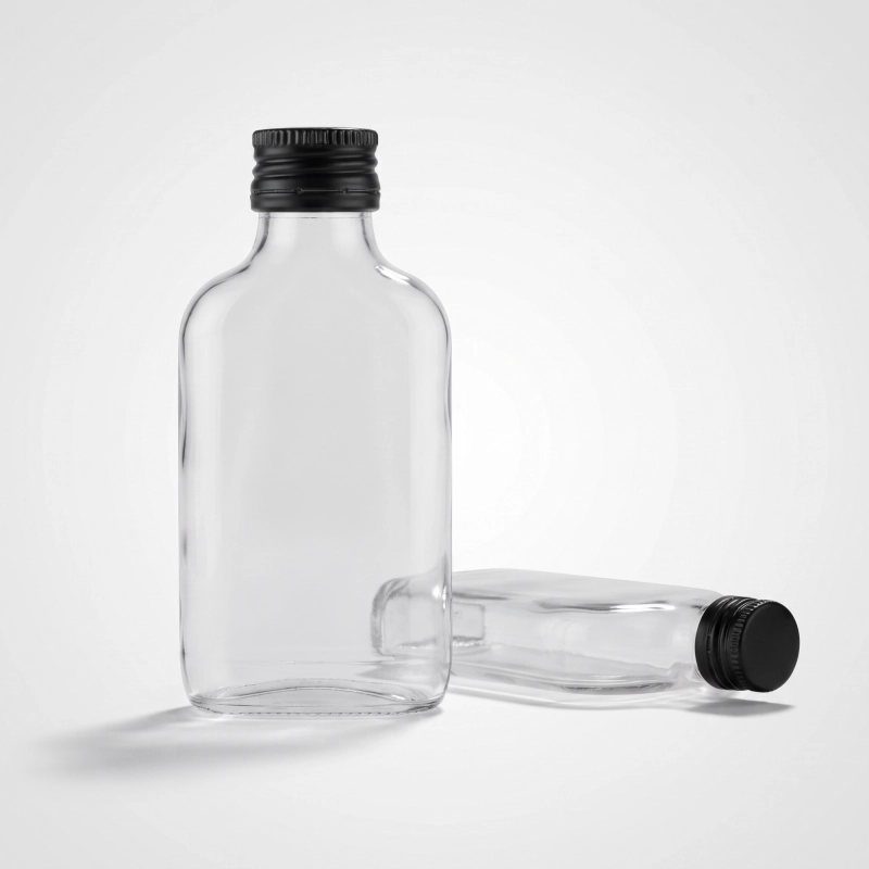 Pusta piersiówka ze szkła bezbarwnego 100 ml z zakrętkami obok, wielorazowa butelka na nalewki